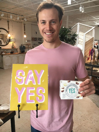 Man holding a "SAY YES" sign and a box of YES Bar macadamia chocolate.