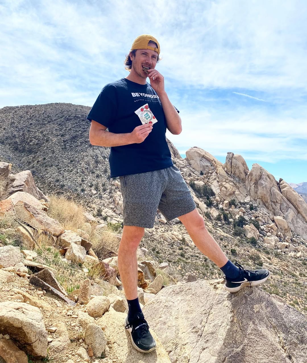 Man enjoying a YES Bar on a rocky hiking trail with mountains under a clear blue sky.