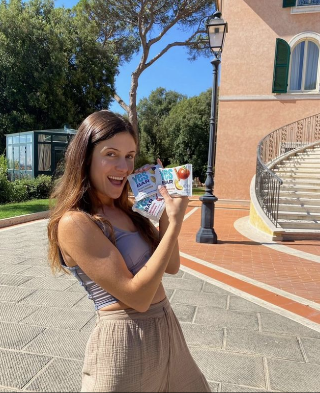 A smiling woman with long brown hair holds three YES Bar snack bars in her hands.