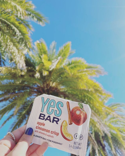 Holding an apple cinnamon crisp ‘YES Bar’ with a palm tree and clear blue sky in the background.
