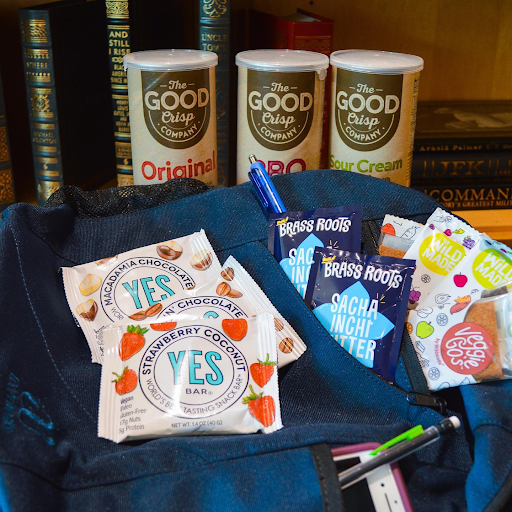 A gift-ready blue tote bag with YES Bars and The Good Crisp Company chips, displayed in front of a modern bookshelf.