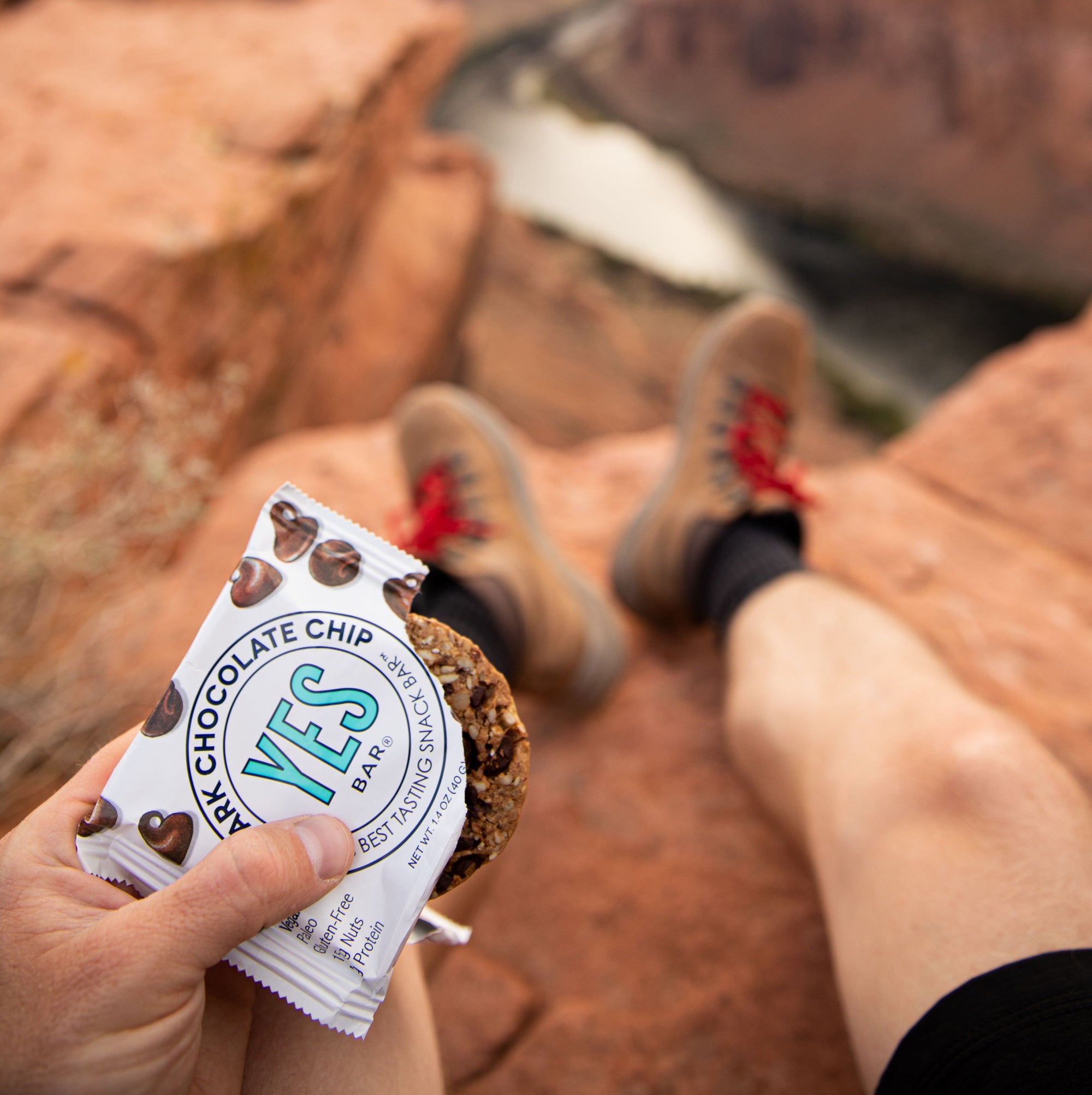 Person on a cliff overlooking a river canyon holding a partially unwrapped Dark Chocolate Chip YES Bar.