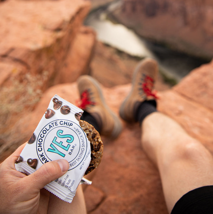 Person on a cliff overlooking a river canyon holding a partially unwrapped Dark Chocolate Chip YES Bar.