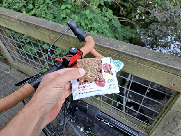 Hand holding a Macadamia Chocolate YES Bar outdoors, with a bicycle handlebar and wood railing suggesting active snacking.