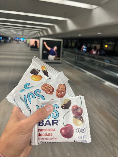 A hand holds three YES Bar snack bars with a blurred airport terminal in the background.