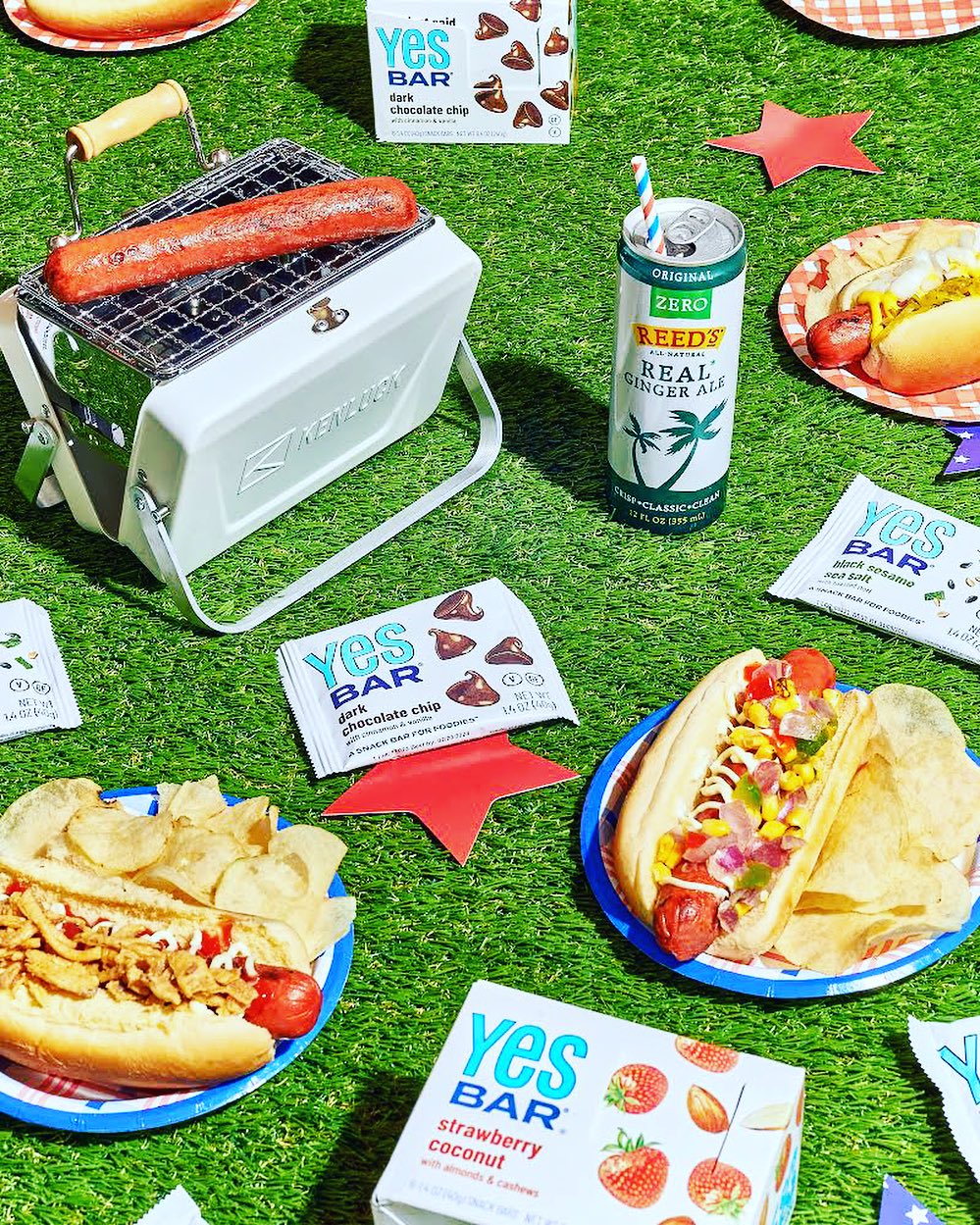 Summer picnic setup with YES Bars, hot dogs on a grill, and snacks displayed on a green lawn.