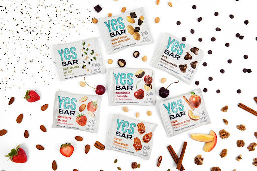 "Seven YES Bar flavors displayed with their ingredients: peanuts, chocolate, almonds, strawberries, cherries, and apples."